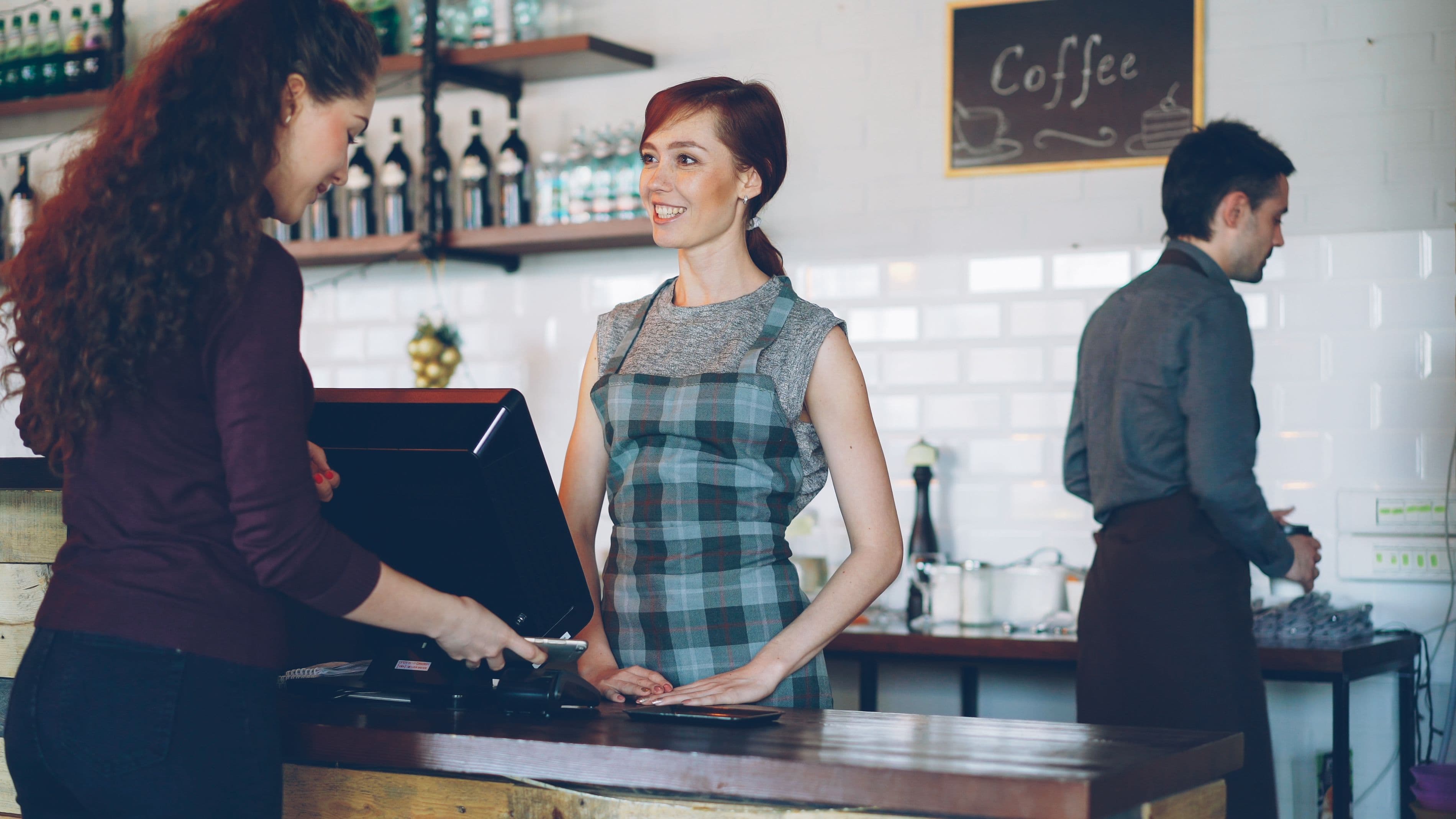A smiling staff member completes a customer's order at the counter while a second employee works independently in the background of a modern cafe.