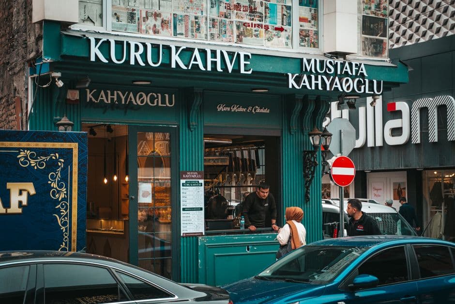 A shopkeeper serves a customer at the counter of a small neighborhood Turkish coffee shop on a busy street.