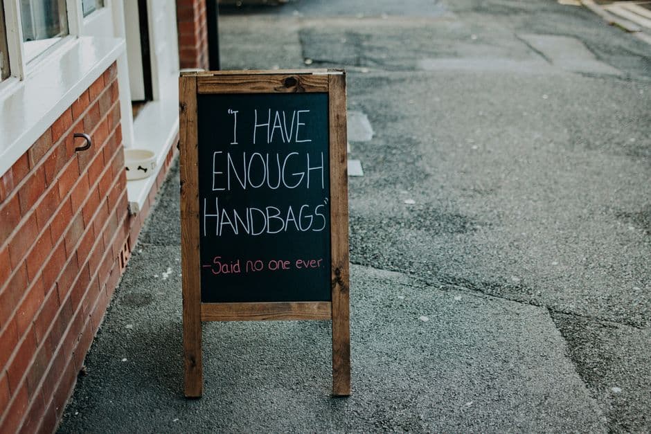 A wooden sandwich-board chalkboard outside a shop reading 'I have enough handbags - said no one ever', an example of a witty tagline.