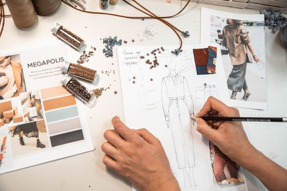 A designer's hand sketches a figure beside a moodboard with a color palette, fabric swatches and beads, illustrating brand identity work beyond a logo.