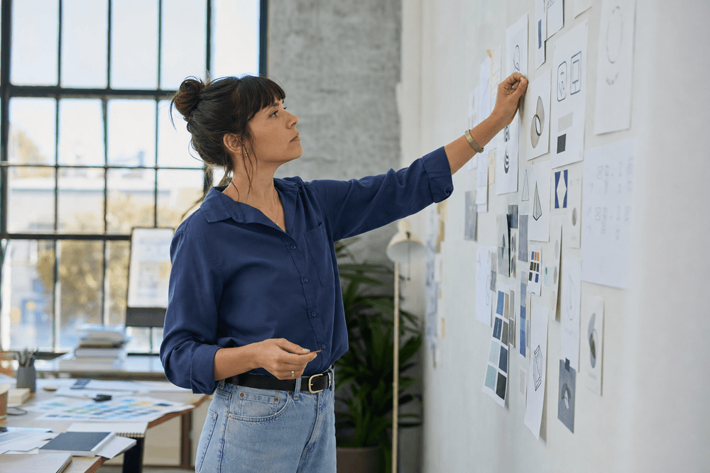 A graphic-studio owner pinning logo sketches and brand swatches to a wall, stepping back to assess.
