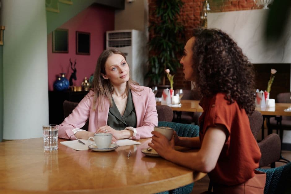 Two women in a warm cafe leaning into an attentive conversation over coffee, with a notepad and pen on the table for taking notes.