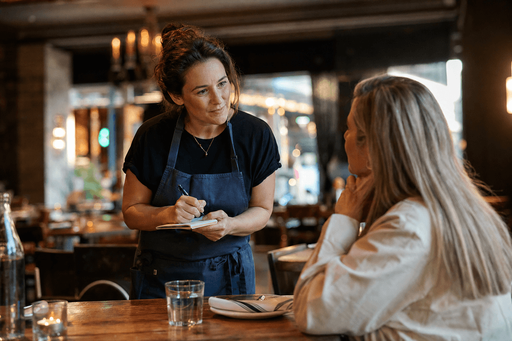A restaurant owner chatting with a regular customer at their table, listening with a notepad.