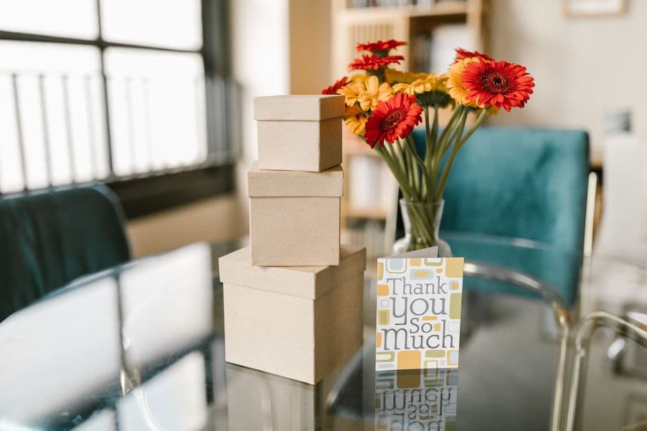 A stack of kraft gift boxes beside fresh red and orange gerbera flowers and a printed 'Thank you so much' card on a glass table.