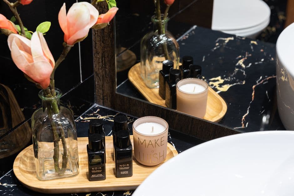 An elegant dark-marble vanity styled with pink magnolia stems, minimalist black amenity bottles on a wooden tray and a lit candle.