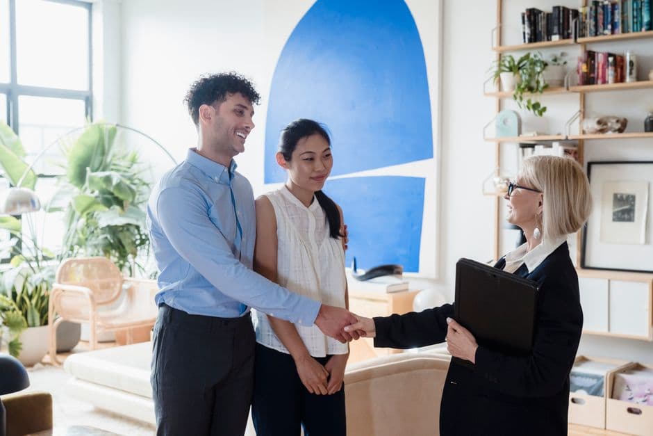 A smiling advisor shakes hands to welcome a young couple in a bright, plant-filled living room with blue abstract wall art.
