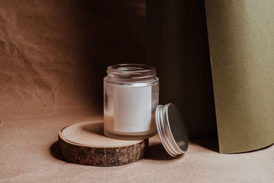 A minimalist frosted-glass candle with a blank label rests on a round wooden slice against a soft earthy backdrop, lit like a premium product.