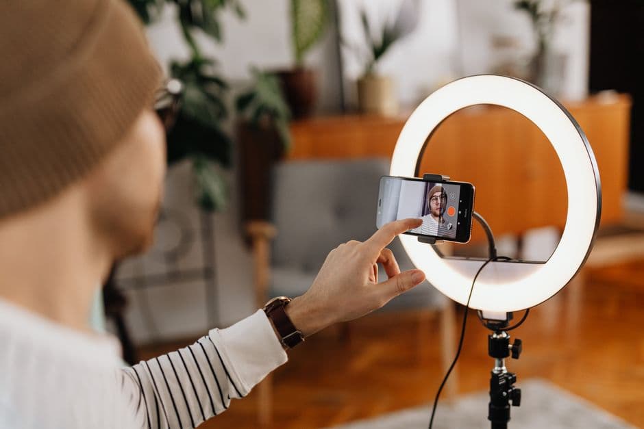 An over-the-shoulder view of a person adjusting a smartphone mounted on a tripod with a ring light to film content in a cozy home studio.