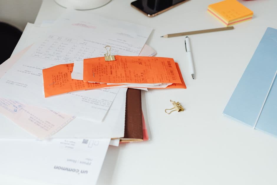 A cluttered white desk covered with loose paper receipts, invoices, sticky notes and pens, evoking repetitive back-office admin work.