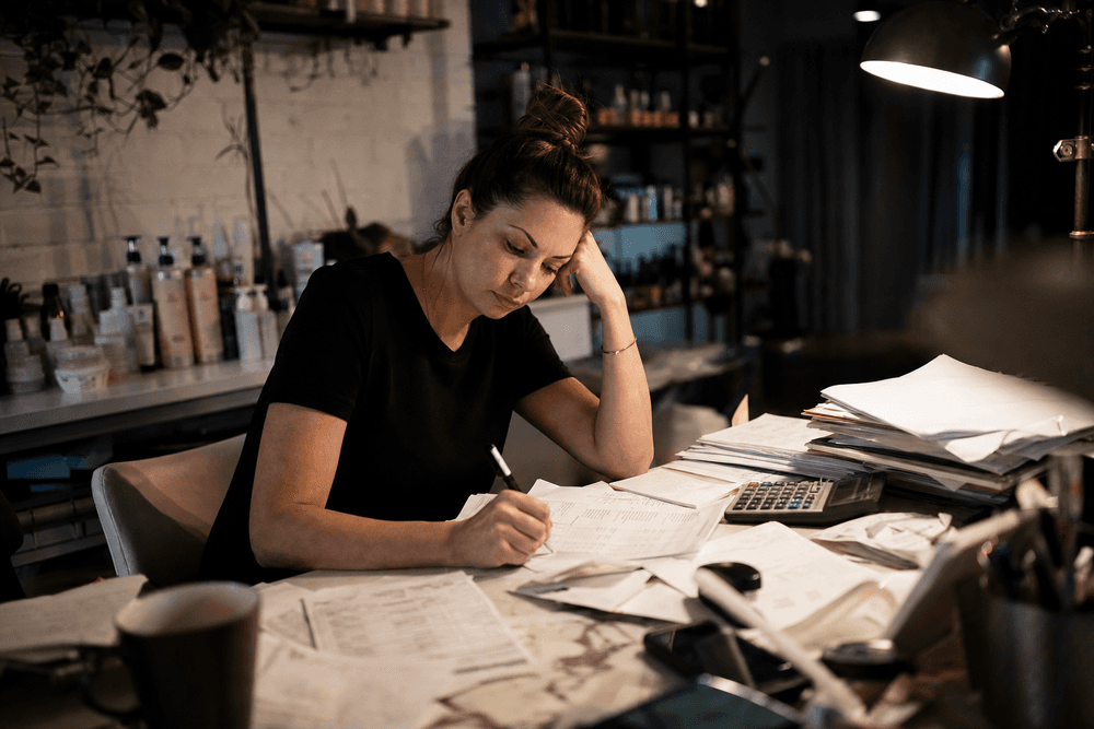 A salon owner buried in a stack of paper invoices and a calculator at a cluttered back-office desk.